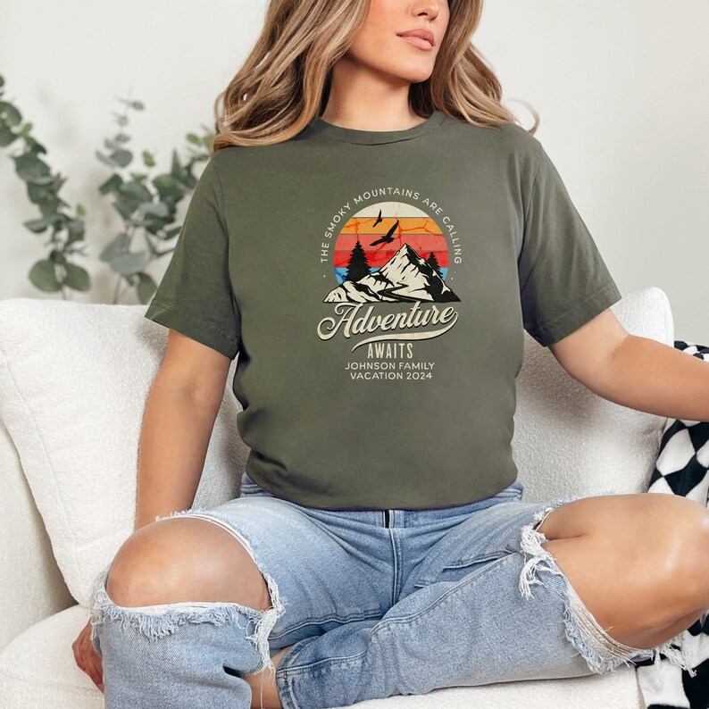 May include: Olive green t-shirt featuring a mountain graphic with the text "The Smoky Mountains are Calling, Adventure Awaits, Johnson Family Vacation 2024". The design includes a sunset, birds, and mountains.