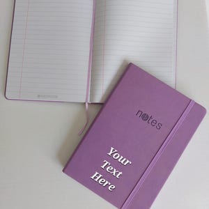 May include: A purple notebook with a ribbon closure and the word "notes" embossed on the cover. The notebook has a blank page for personalisation with the text "Your Text Here".