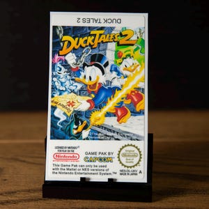 May include: A rectangular DuckTales 2 video game cartridge for the Nintendo Entertainment System. The label features Scrooge McDuck, Huey, Dewey, and Louie in a colourful illustration. Text includes the game title, Capcom branding, and Nintendo seal.