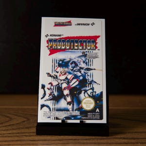 Może przedstawiać: Prostokątna kaseta z grą wideo na system Nintendo Entertainment System z tytułem PROBOTECTOR zapisanym niebieskimi i czerwonymi literami. Etykieta przedstawia dwie futurystyczne postacie w biało-czerwonych zbrojach na ciemnym tle.