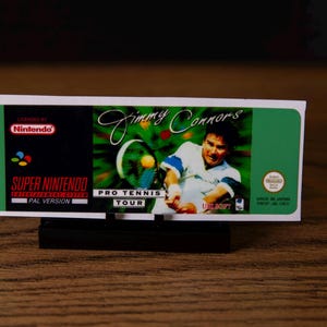 Peut inclure: Cartouche de jeu Super Nintendo pour "Jimmy Connors Pro Tennis Tour". L'étiquette présente Jimmy Connors jouant au tennis, avec le logo Nintendo et le titre du jeu. Version PAL, dimensions en cm.