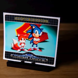 May include: A retro video game cartridge featuring Sonic the Hedgehog and Tails, with the text "SONIC THE HEDGEHOG 2" and "MEGA DRIVE". The characters are set against a teal background with a red and checkered design.