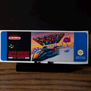 May include: A rectangular F-Zero video game label for the Super Nintendo Entertainment System. The label features a blue border, the Nintendo logo, and a graphic of a futuristic racing vehicle. Text includes Licensed by Nintendo, Super Nintendo Entertainment System, PAL Version, and Made in Japan.