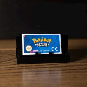 May include: A rectangular video game cartridge for a handheld console featuring a blue label with the text Pokémon Sapphire Version, a Nintendo seal, a CE mark, and the product code AGB-AXPP-EUR, displayed on a black plastic stand.