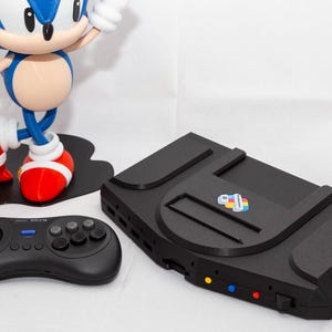 May include: A black video game console with a central cartridge slot and a small colorful logo, accompanied by a black six-button game controller and a blue, white, and red Sonic the Hedgehog figurine holding a gold ring on a black base.