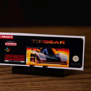 May include: A Super Nintendo game cartridge for Top Gear. The cartridge has a black background with the game title in orange and an image of a race car. The cartridge is licensed by Nintendo and made in Japan.