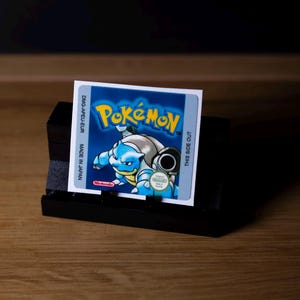 May include: A Pokemon Game Boy cartridge label with a blue Blastoise design on a blue background, and the word "Pokemon" in yellow. The label is on a black stand, with text that reads "Made in Japan" and "This Side Out."