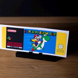 May include: A Super Mario World video game cartridge label. The label features Mario riding Yoshi on a blue background. The Nintendo logo is visible, along with the text "SUPER MARIO WORLD" and "PAL VERSION".