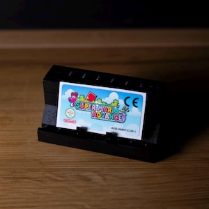 May include: A black Nintendo Game Boy Advance cartridge holder with a Super Mario Advance game cartridge. The cartridge has a colorful label with the game title and Nintendo logo. The holder is on a wooden surface.