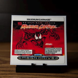 Puede incluir: Una etiqueta cuadrada de cartucho de videojuego para Spider-Man y Venom: Maximum Carnage para Sega Mega Drive. La etiqueta presenta un fondo rojo con una ilustración negra de Carnage, el logotipo de Acclaim y texto blanco sobre licencias.