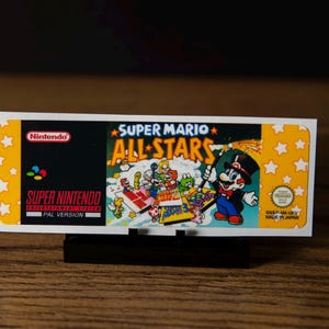 May include: A rectangular Super Mario All-Stars video game label for the Super Nintendo Entertainment System. The label features a black section with the Nintendo logo and a yellow section with white stars. Mario is depicted in a tuxedo and top hat.