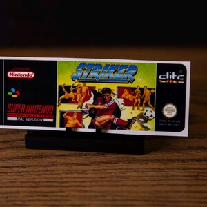 Puede incluir: Un cartucho de juego de Super Nintendo para el juego "Striker". El cartucho presenta una colorida ilustración de un jugador de fútbol pateando un balón, con el título del juego en letras azules en negrita. El cartucho está etiquetado como "PAL Version".