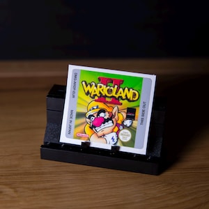 以下が含まれることがあります： 黒いスタンドに表示されたWario Land II Game Boyカートリッジ。カートリッジには、ゲームのタイトルとワリオの漫画の画像が描かれたカラフルなラベルが付いています。ラベルは緑、黄色、白の色をしています。