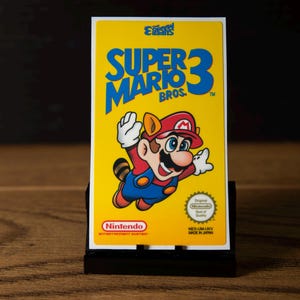May include: A rectangular Super Mario Bros. 3 video game cartridge with a bright yellow label. The label features the blue title text, a graphic of Mario in a raccoon suit, the Nintendo logo, and an Original Nintendo Seal of Quality emblem.