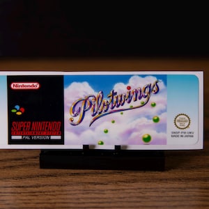 Puede incluir: Un cartucho de juego de Super Nintendo para Pilotwings. La etiqueta presenta el logotipo de Nintendo, el título del juego en una fuente estilizada y un fondo temático de cielo con nubes y orbes verdes. El cartucho está etiquetado como "PAL VERSION".