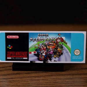 Puede incluir: Un cartucho de juego Super Nintendo Super Mario Kart. La carátula muestra a Mario y otros personajes compitiendo en una pista. El logotipo de Nintendo es visible a la izquierda, con el texto "PAL VERSION". El cartucho está sobre un soporte negro.