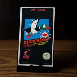 Op de afbeelding: Een rechthoekige Duck Hunt videogamecartridge voor het Nintendo Entertainment System. Het zwarte label toont een gepixelde eend in vlucht tegen een blauwe achtergrond, het rode Duck Hunt logo, een lichtpistool icoon en het Nintendo kwaliteitszegel.
