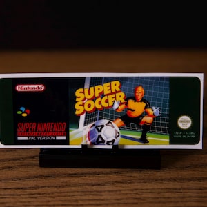 Puede incluir: Cartucho de juego de Super Nintendo para Super Soccer. La etiqueta presenta el logotipo de Nintendo, el título del juego en amarillo y una ilustración de un portero de fútbol. El cartucho es negro y está sobre un soporte negro.
