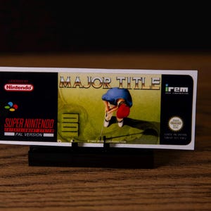 Pode incluir: Um cartucho de jogo Super Nintendo para "Major Title" é exibido. O rótulo apresenta o logotipo da Nintendo, o título do jogo e um gráfico de golfista. O cartucho é a versão PAL e fabricado no Japão.