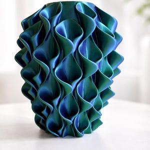 Può includere: Un vaso decorativo con un design unico e strutturato. Il vaso è una miscela di verde acqua e blu, con un motivo a onde e stratificato. È un pezzo d'arredo per la casa di grande effetto.