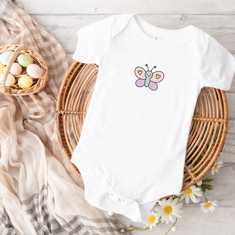 Puede incluir: Un body blanco para beb&eacute; con una mariposa rosa y morada con corazones amarillos en la parte delantera.