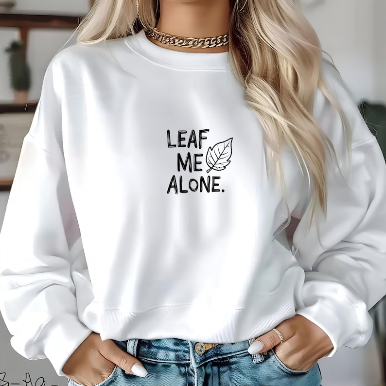 Puede incluir: Sudadera blanca con un gr&aacute;fico negro que dice "LEAF ME ALONE." y una peque&ntilde;a ilustraci&oacute;n de hoja.