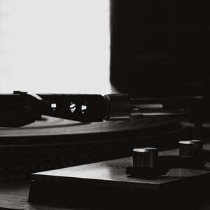 May include: Monochrome image of a vintage record player. The turntable's arm is positioned over a vinyl record. The control panel features silver knobs. The background is softly lit, with a window and blinds visible.