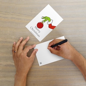 May include: Two white Christmas cards with the Grinch holding a red ornament. One card has the text "Merry Christmas" and "From Whoville". A person is writing on the other card with a black pen. The background is a light brown wooden surface.
