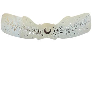 May include: A cream-coloured, wing-shaped decorative piece with a celestial design. The design includes a crescent moon, stars, and floral elements. The piece is cut out with intricate details, creating a delicate and ethereal aesthetic.