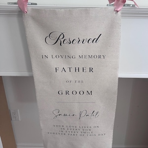 Peut inclure: Une bannière rectangulaire en tissu beige avec un texte imprimé en noir indiquant Reserved IN LOVING MEMORY FATHER OF THE GROOM Samir Patel YOUR LOVE LIVES ON IN EVERY VOW IN EVERY SMILE FOREVER PART OF THIS DAY. La bannière a des nœuds en ruban rose.