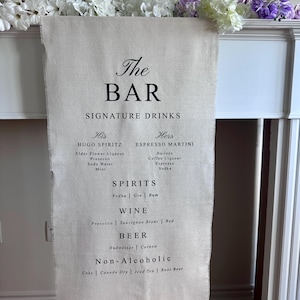 Op de afbeelding: Een verticale beige stoffen banner met zwarte gedrukte tekst die The BAR SIGNATURE DRINKS leest, met een lijst van Hugo Spritz, Espresso Martini, Spirits, Wine, Beer en Non-Alcoholic opties met hun respectievelijke ingrediënten en dranksoorten.