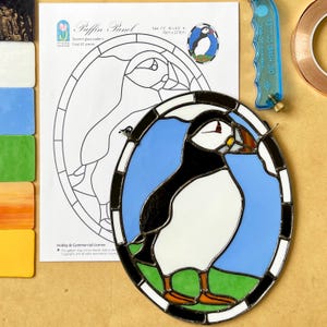 Könnte beinhalten: Ein Buntglas-Papageientaucher-Panel mit einem schwarzen, weißen und blauen Design. Das Panel ist oval und zeigt eine detaillierte Darstellung eines Papageientauchers. Größe: 19 cm B x 22,9 cm H.
