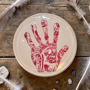 May include: A round, off-white ceramic plate featuring a red graphic print of a handprint. Within the shape of the hand, there is a detailed illustration of a person wearing a traditional headdress. The plate has a smooth, glossy finish.