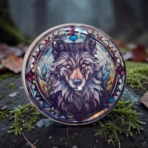Könnte beinhalten: Ein kreisförmiger Glaskabochon mit einer detaillierten Illustration eines Wolfes mit bernsteinfarbenen Augen vor einem Hintergrund im Buntglasstil mit blauen, grünen und roten geometrischen Mustern und Blattmotiven, eingefasst in einen klaren Rahmen.