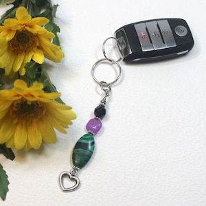 May include: A green, purple, and black beaded keychain with a silver heart charm. The keychain is attached to a silver key ring.