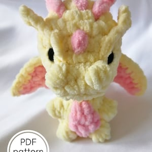 Peut inclure: Un jouet en peluche de dragon crocheté en fil jaune et rose. Le dragon a un petit nez rose et des cornes roses. Le jouet est posé sur une surface blanche. Modèle PDF uniquement.