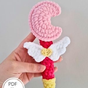 Peut inclure: Une baguette crochetée à la main avec un croissant de lune rose, des ailes blanches et un cœur jaune. Le manche de la baguette est rouge avec une pointe jaune. Le texte "PDF pattern only" est dans un cercle blanc.