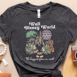 May include: Dark gray t-shirt with a Walt Disney World graphic and the text "The happiest place on earth." The image includes a straw hat, passport, camera, and other travel accessories.