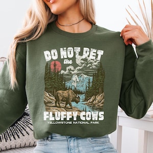 Puede incluir: Sudadera verde militar con un gr&aacute;fico que dice "Do Not Pet the Fluffy Cows" sobre un paisaje del Parque Nacional de Yellowstone. El dise&ntilde;o presenta un bisonte, monta&ntilde;as, &aacute;rboles y un r&iacute;o. Cuello redondo.