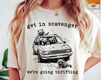 May include: Cream-colored t-shirt with a black graphic of raccoons in a car, with the text "get in scavenger we're going thrifting". The shirt is a casual style, suitable for everyday wear.