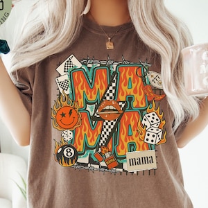 May include: Brown t-shirt with a graphic design featuring the word "MAMA" in a flame-like font, surrounded by dice, playing cards, and a smiley face. The shirt has a vintage, worn look.