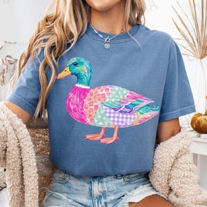 May include: A blue jean colored short-sleeve t-shirt featuring a graphic of a mallard duck with a colorful patchwork pattern on its body, including pink, purple, and teal sections with star and plaid prints, and a bright orange beak and feet.
