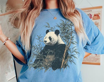 May include: A light blue t-shirt featuring a panda bear illustration surrounded by bamboo. The panda is black and white, and the bamboo is green. The text "Blue Jean" is written in cursive in the upper right corner.