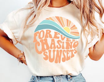 May include: Cream-colored t-shirt with the text "FOREVER CHASING SUNSETS" in orange, with a graphic of a sunset and waves in blue and orange. The shirt is paired with light blue jeans.