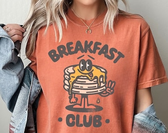 May include: Orange-colored t-shirt with the words "BREAKFAST CLUB" and a cartoon pancake character. The pancake has eyes, a peace sign, and syrup dripping down. The shirt is a vintage style.