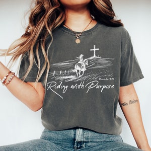 May include: A gray t-shirt with a white graphic of a person riding a horse towards a cross. The text on the shirt reads 'Proverbs 19:21' and 'Riding with Purpose'. The shirt is being worn by a person wearing blue jeans and white shoes.