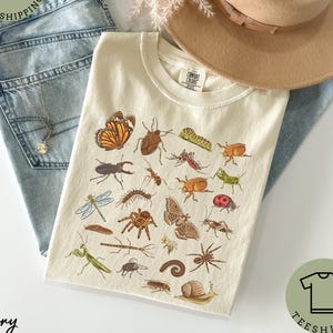 May include: Ivory-colored t-shirt featuring a detailed illustration of various insects, including a butterfly, beetle, and spider. The shirt is paired with light blue jeans and a tan hat. The text "Comfort Colors" is visible on the shirt's tag.