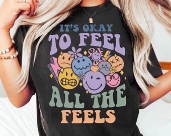 May include: Black t-shirt with the text "IT'S OKAY TO FEEL ALL THE FEELS" in a retro font. The design includes various smiley faces, flowers, and lightning bolts in pastel colors. The brand name "Tee Shirtophia" is at the bottom.