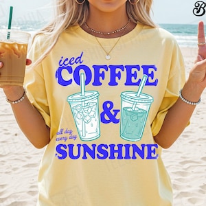 May include: Yellow t-shirt with the words "iced COFFEE & SUNSHINE" in blue. The shirt features illustrations of iced coffee drinks. The phrase "all day every day" is also printed on the shirt. The person is holding an iced coffee drink.