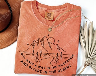 Puede incluir: Una camiseta Comfort Colors de color coral con un gráfico negro de montaña y río. El texto dice "I will make a way in the wilderness and rivers in the desert." Un collar dorado y un sombrero marrón también están en la imagen.